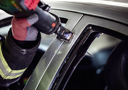 Person som använder ett verktyg på en bils dörrkaross, iklädd skyddsutrustning med reflexdetaljer.