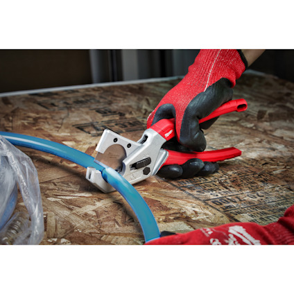 Röd- och vitfärgad skärverktyg klipper ett blått rör på en arbetsskiva. Handen bär en svart och röd handske.