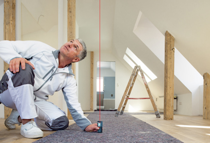 Man använder en laseravståndsmätare på golvet i ett renoverat rum med träbjälkar och ett stege i bakgrunden.